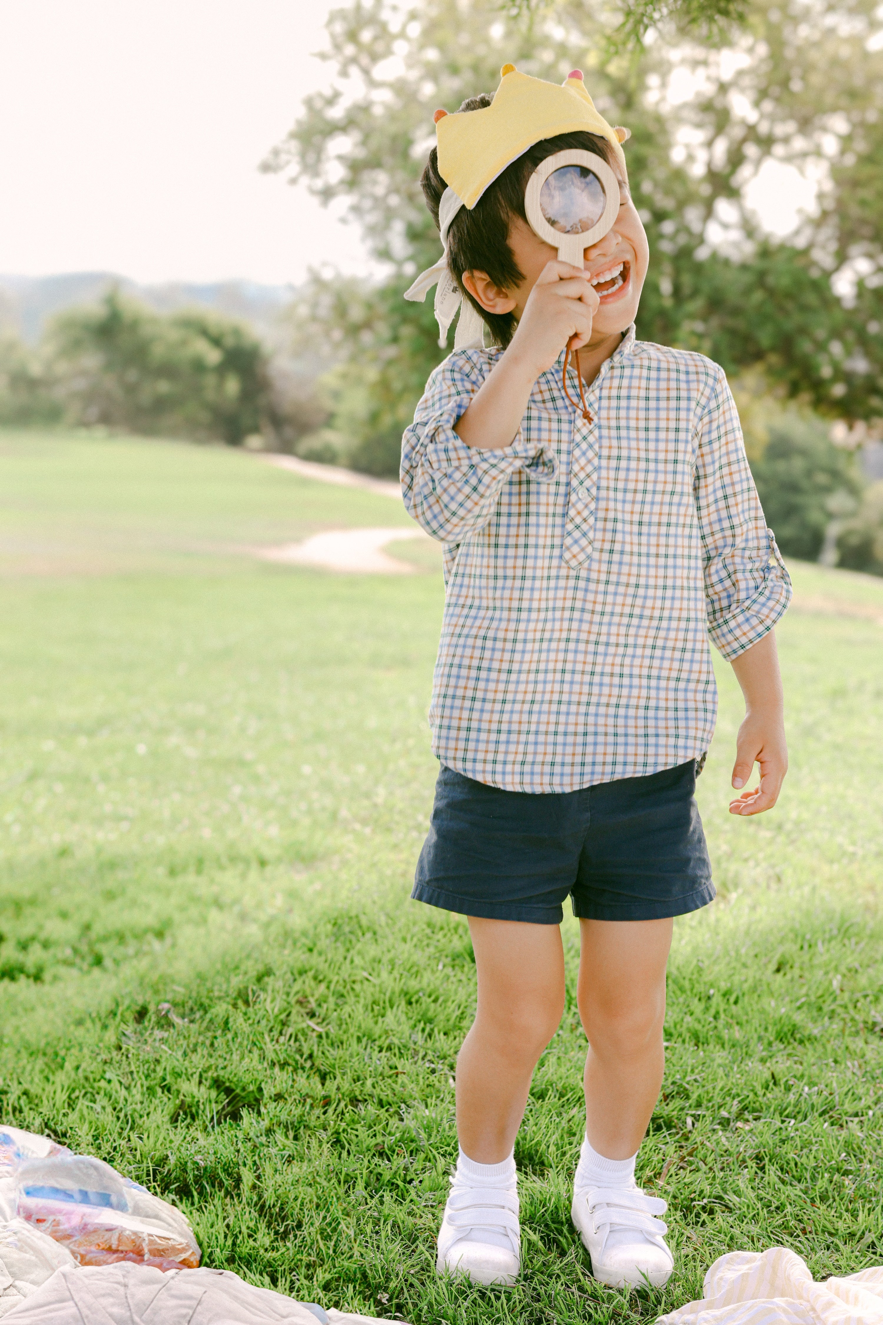 Child playing outdoors with a magnifying glass and crown, standing on grass.
