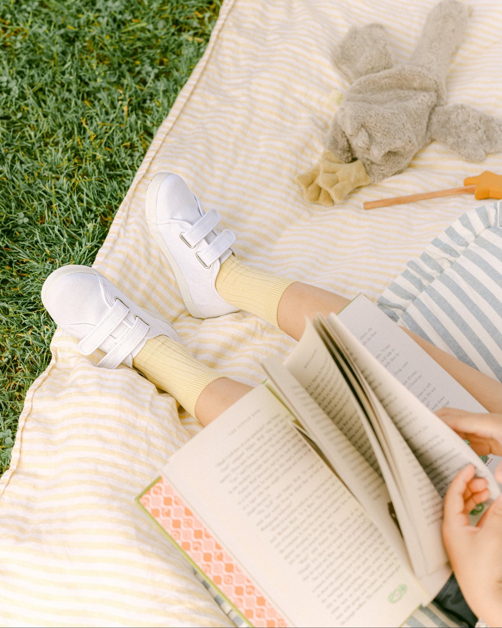 Person reading a book on a blanket outdoors with a teddy bear and straw in the background